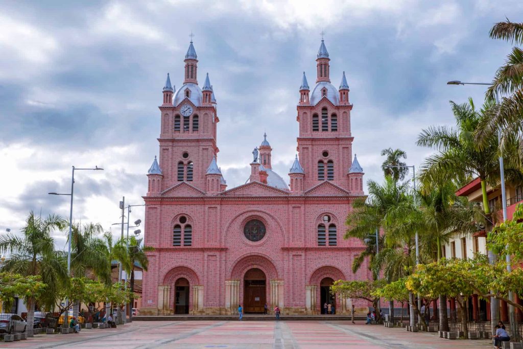 Fachada principal de la Basílica del Señor de los Milagros en Buga, Colombia, con sus dos torres simétricas de color rosado, detalles arquitectónicos en ladrillo y una plaza amplia con palmeras al frente.